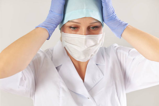 Πορτρέτο ενός γιατρού σε ένα ιατρικό παλτό, μάσκα και γάντια σε λευκό φόντο. Μια γυναίκα στο κεφάλι με τα χέρια. - Φωτογραφία, εικόνα