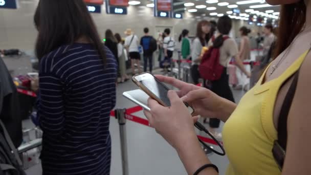 4K Asian turystka sprawdza telefon, jak czeka na check-in Liczniki na międzynarodowym lotnisku. Ludzie korzystający ze smartfona zrobić szybkie wiadomości tekstowe z przyjaciółmi w sieci społecznej na wakacje podróży-Dan - Materiał filmowy, wideo