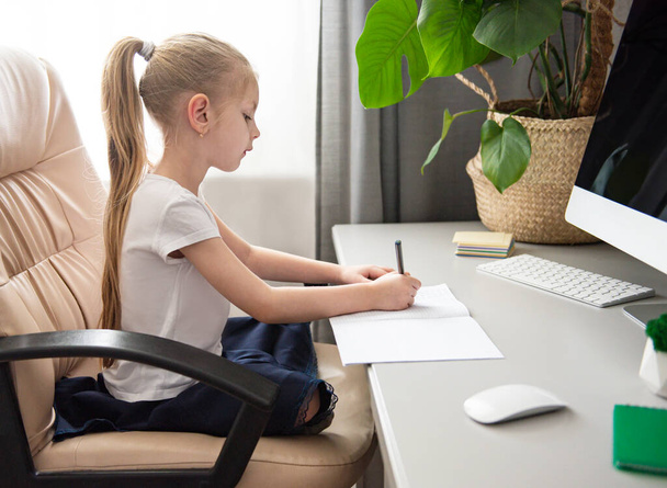 Оставайтесь дома для предотвращения пандемии коронавируса. Маленькая девочка с светлыми волосами сидит за столом и делает домашнее задание
.  - Фото, изображение