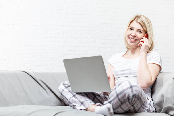 Smiling blonde woman is sitting on the couch and working online. Remote work during the coronavirus pandemic. Space for text. - Photo, image