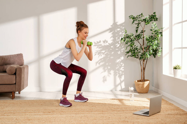 Вид сбоку активной молодой женщины в спортивном костюме, выполняющей упражнения на приседание с гантелями и смотрящей видео на ноутбуке во время фитнес-тренировки в хоме
 - Фото, изображение