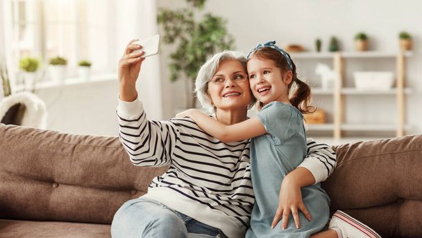 Mujer mayor sonriendo y tomando selfie con chica alegre mientras se sienta en el sofá en la acogedora sala de estar en hom - Foto, imagen