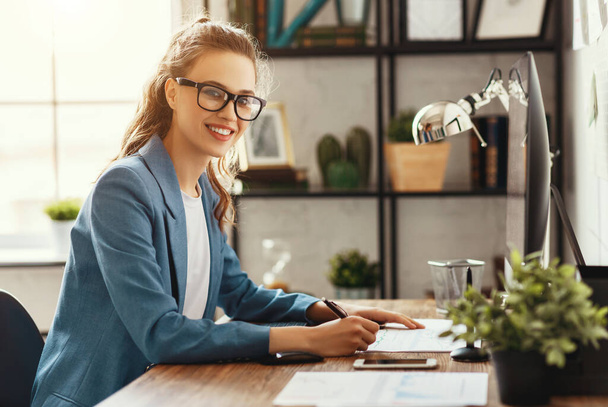 Widok z boku pozytywnej młodej kobiety przedsiębiorcy w formalnym stroju i okularach patrząc w aparat fotograficzny siedząc przy stole i robiąc notatki na papierach w nowoczesnym miejscu pracy - Zdjęcie, obraz