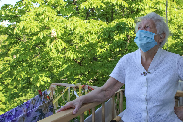 Koncepcja Covid-19, zdrowie, bezpieczeństwo i pandemia - starsza samotna kobieta nosząca ochronną maskę medyczną stojąca na balkonie w domu dla ochrony przed wirusem - Zdjęcie, obraz