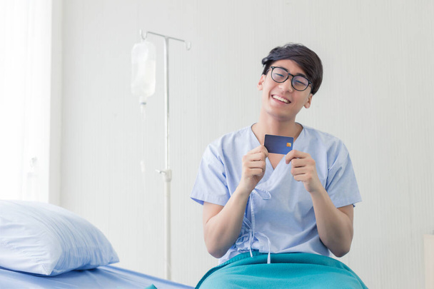 Junger asiatischer Teenager mit Kreditkarte sitzt mit Smiley-Gesicht auf dem Bett im Krankenhaus. - Foto, Bild