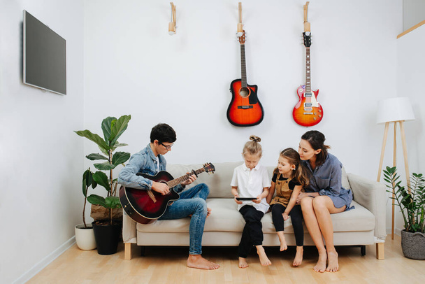 Family spending time on a sofa in the living room, eash doing his or her own thing. Dad playing guitar, mom watching kids play. - Foto, Imagen