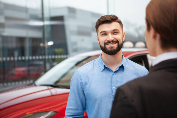 Vrolijke knappe man glimlachend vrolijk tijdens het praten met auto dealer manager, het kopen van nieuwe auto, kopiëren ruimte - Foto, afbeelding