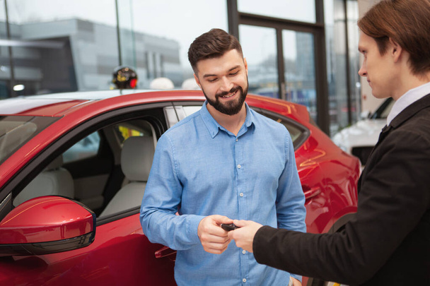 Charming bearded man receiving car keys from the salesman after buying a new automobile, copy space. Deal, contract, rental concept - Foto, imagen