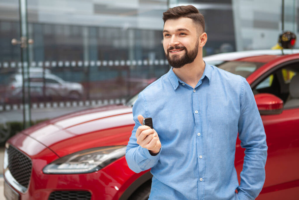 Veselý pohledný muž se usmívá, dívá se jinam, drží klíče od svého nového auta v prodejně aut, kopíruje prostor - Fotografie, Obrázek