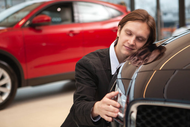 Young handsome man smiling with his eyes closed, embracing new car at the dealership - Foto, Imagem