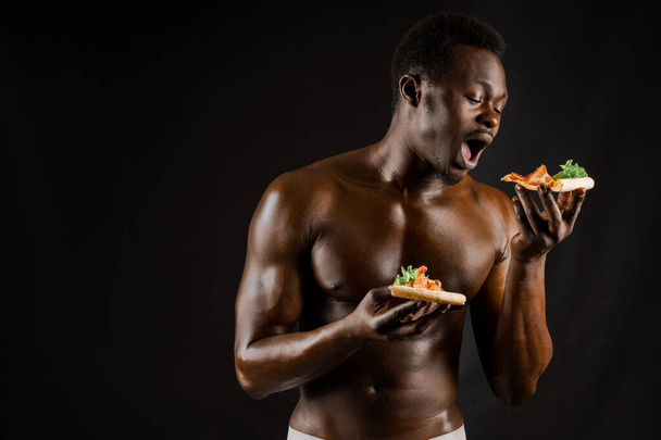Black handsome naked man eats a pizza. Safety food delivery for people who stays at home. Sexy african man shows 2 pieces of pizza in his hands. Tasty food from pizzeria restaurant - Foto, Imagem