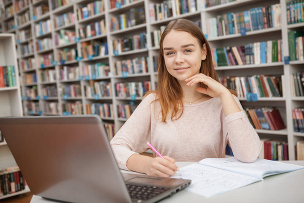 Fröhliches Teenager-Mädchen lächelt in die Kamera, während sie an ihrem Laptop in der College-Bibliothek arbeitet, Kopierraum. E-Learning, Bildungskonzept - Foto, Bild