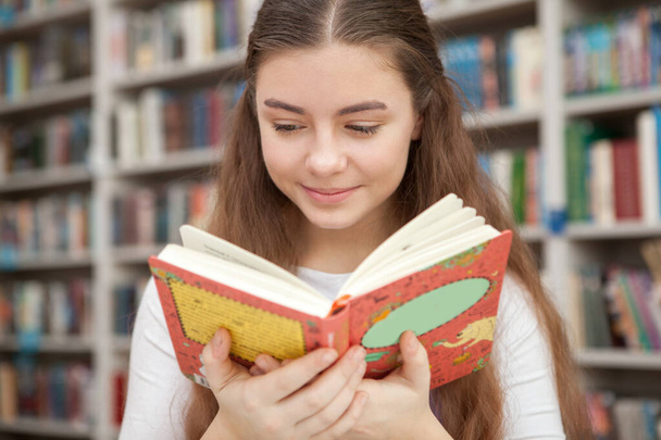 Крупный план очаровательной девушки-подростка, улыбающейся, читающей книгу в местной библиотеке. Воображение, хобби, концепция расслабления
 - Фото, изображение