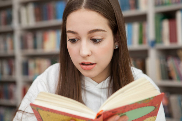 Közelkép egy tinédzser lányról, aki túlterheltnek tűnik, miközben könyvet olvas a könyvtárban. - Fotó, kép