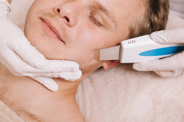 Recortado cerca de un joven guapo relajándose mientras dermatólogo usando un dispositivo de limpieza por ultrasonido en su piel
 - Foto, imagen