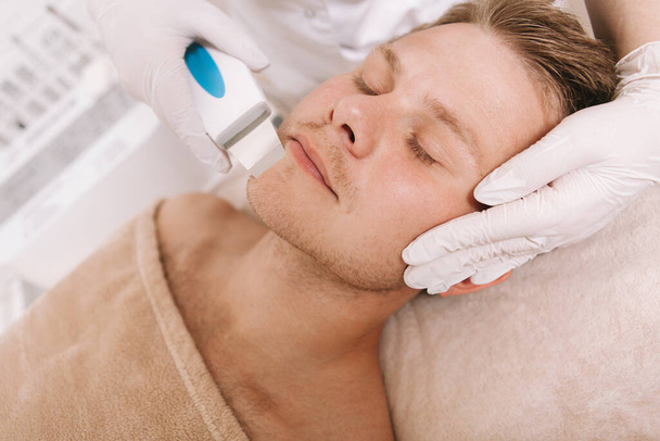 Blisko atrakcyjnego mężczyzny leczącego skórę twarzy przez kosmetologa. Kosmetyk stosujący ultradźwiękowe urządzenie oczyszczające u męskiego klienta - Zdjęcie, obraz
