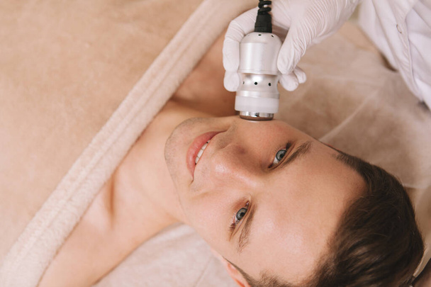 Top uitzicht close-up van een knappe vrolijke man met huid verstevigende gezichtsbehandeling bij cosmetologie salon - Foto, afbeelding