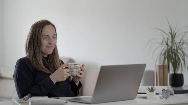 Smiling young woman freelancer using laptop computer, typing message, drinking coffee, distance online worker concept. - 写真・画像