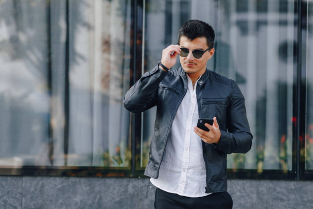 young stylish guy in glasses in black leather jacket with phone on simple glass background - Photo, Image