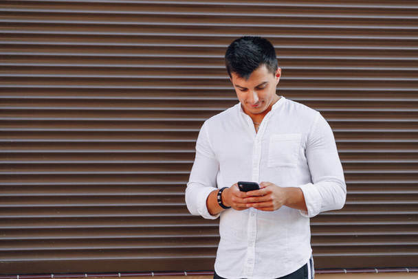 νεαρός κομψός τύπος σε πουκάμισο πληκτρολογώντας στο τηλέφωνο σε απλό καφέ φόντο - Φωτογραφία, εικόνα