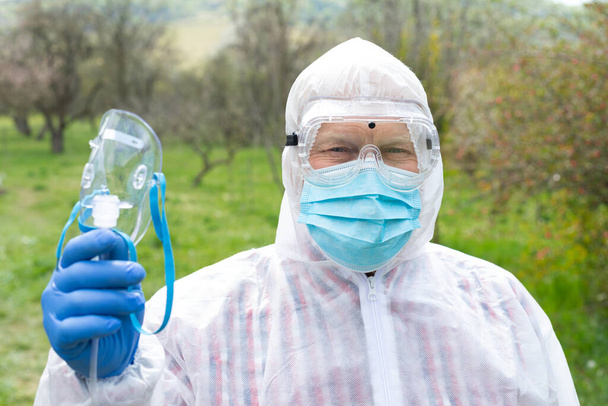 Portrait of senior man wearing hazmat suit, glasses, mask and gloves is holding an oxygen mask outdoors - Foto, Imagem