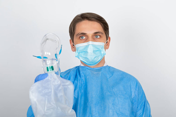 Young male doctor wearing protective uniform is holding an oxygen mask on isolated background - Photo, image