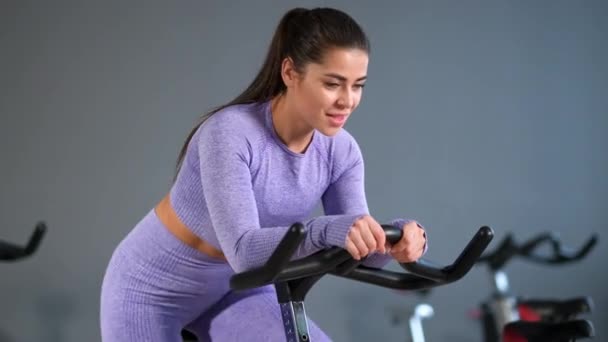 Una bella giovane donna bruna atletica in abbigliamento sportivo si allena su un sycle in palestra contro il muro grigio. - Filmati, video