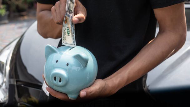 Ein junger Mann steht vor einem Auto und steckt Geld in ein Sparschwein. Sparen, um ein Haus zu kaufen, Häusersparnisse, Kredite Markt concep - Foto, Bild