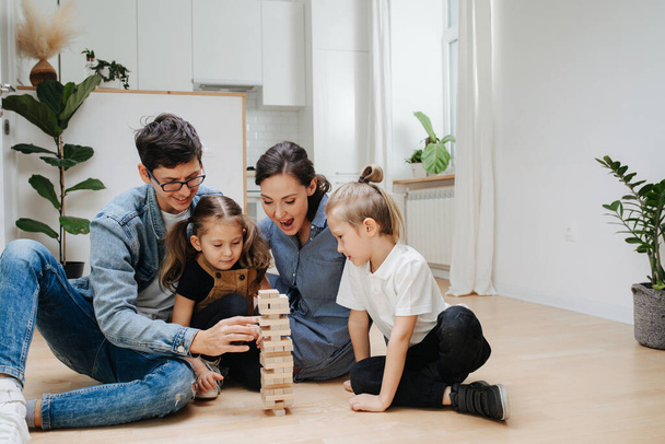 Mom, dad playing with kids on the kitchen floor, building tower from wooden blocks, taking tiles in turn from underneath untill it falls. Spending time with kids on isolation. - Фото, изображение