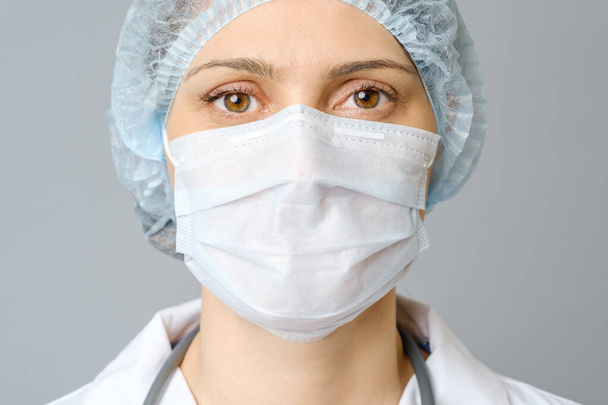 Portrait of young female doctor in protective medical mask on her face and cap on her head. Isolated on gray background - Foto, imagen