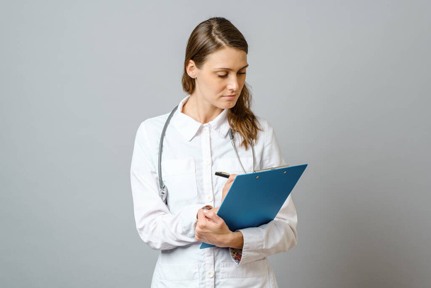 Young woman physician with stethoscope prescribing treatment to patient. Female doctor with pen writing recipe on clipboard. Isolated on gray - Fotoğraf, Görsel