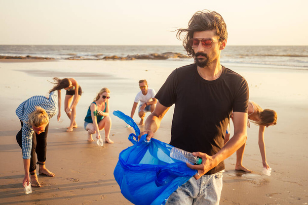 handsome indian man in the foreground collecting plastic from sand into the bag , his mixed race friends picking waste on background in Goa India beach - Foto, afbeelding