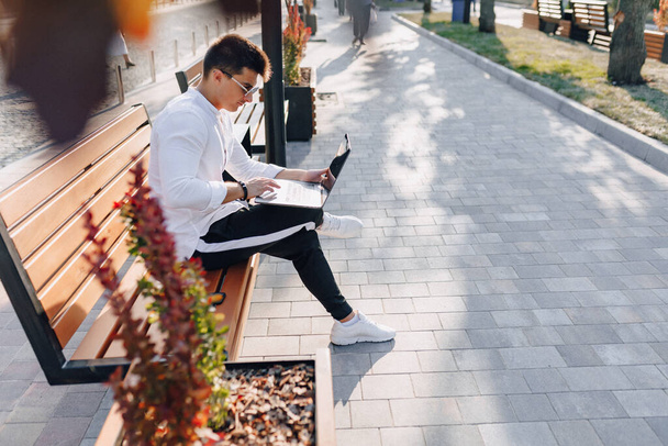 young stylish guy in white shirt with phone and notebook works on bench on sunny warm day outdoors, freelance - Фото, изображение