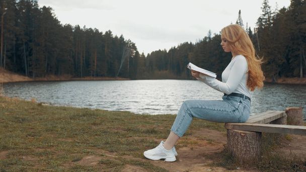Beautiful young blonde woman sitting and reading book in a park near the lake. Side view - Zdjęcie, obraz