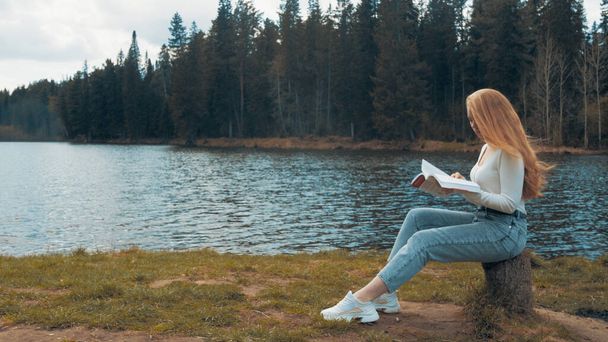 Bella giovane donna bionda seduta a leggere un libro in un parco vicino al lago. Vista laterale - Foto, immagini