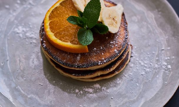 Desayuno casero de panqueques decorados con naranja, plátano, menta y azúcar en polvo sobre fondo negro en plato gris
. - Foto, imagen