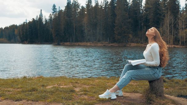 Beautiful young blonde woman sitting and reading book in a park near the lake. Side view - Fotografie, Obrázek