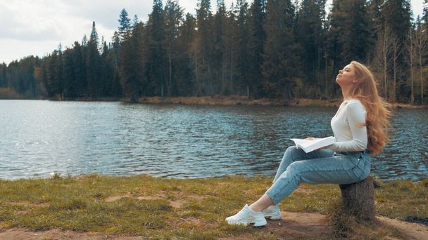 Beautiful young blonde woman sitting and reading book in a park near the lake. Side view - Фото, зображення