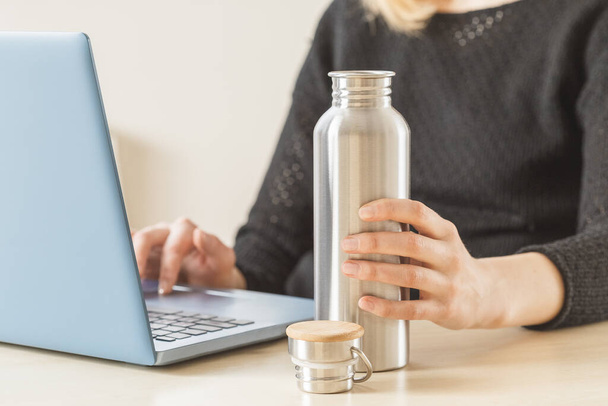 Femme blanche tenant sa bouteille d'eau personnelle en acier inoxydable sur la table de travail. Habitude d'hydratation quotidienne pour rester en bonne santé
 - Photo, image