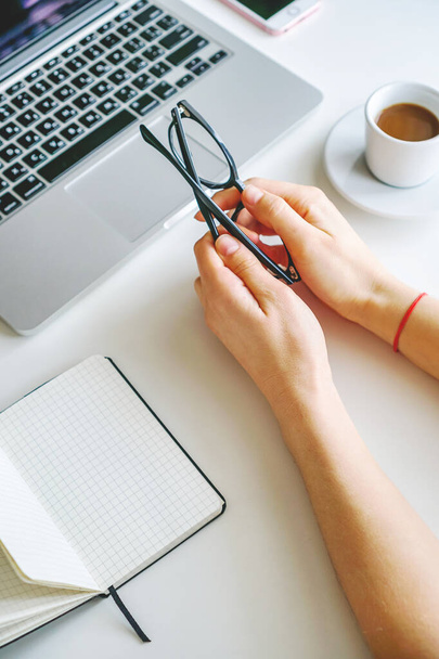girl holds glasses in her hands. nearby is a notebook, there is a coffee mug and a laptop - Photo, Image