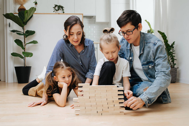 Mom, dad and kids lying on the kitchen floor, building wall from wooden blocks. Spending quality time together on isolation. - Photo, Image