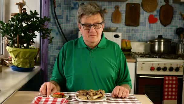 Hongerige oudere volwassen man, gebonden servet om zijn nek, met plezier likken, kijkend naar varkensvlees aan tafel in zijn thuiskeuken. Middelgroot plan. - Video