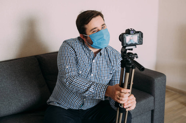 man blogger wearing protective face mask is recording video on video camera. Male blogger streaming about coronavirus pandemic. Stay home for social distancing containment self quarantine - Foto, imagen