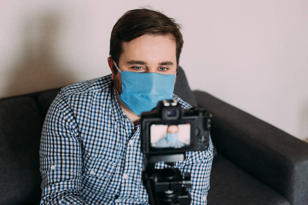 Young man blogger wearing protective face mask is recording video on video camera. Male blogger streaming about coronavirus pandemic. Stay home for social distancing containment self quarantine - Φωτογραφία, εικόνα