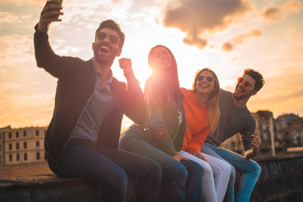 Happy young people taking a selfie sitting on a bench outdoors in the sunset. Real backlight flare. Leisure activities lifestyle concept. - Zdjęcie, obraz