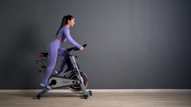Une belle jeune femme brune sportive en vêtements de sport s'entraîne sur un sycle dans la salle de gym contre le mur gris. - Séquence, vidéo