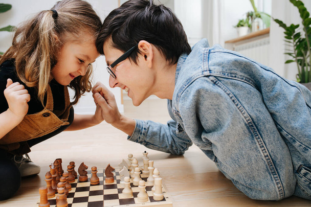Happy father and his little daughter playing chess on a kitchen floor at home. Side view. Struggling over the board, pushing against each other's head and hands. - Fotó, kép