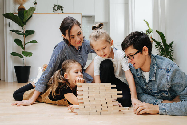 Mom, dad and kids lying on the kitchen floor, building wall from wooden blocks. Spending quality time together on isolation. - Fotoğraf, Görsel