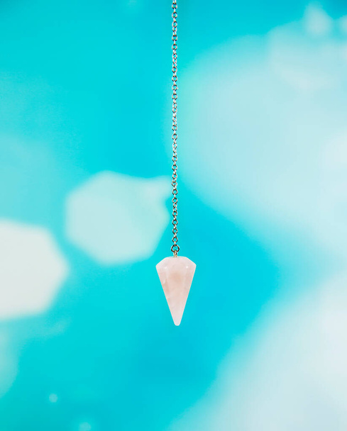 Enchanted Rose Quartz Pendulum point hanging on silver chain, isolated on blue bokeh background in studio. Prediction concept. Meaning:stone of the heart, divine loving energy. - Photo, Image