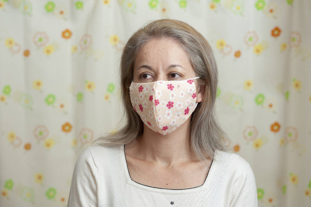 Porträt einer Frau mittleren Alters, die eine selbstgemachte Gesichtsmaske aus Stoff trägt, um sich vor Coronavirus (COVID-19) und anderen Infektionskrankheiten zu schützen. Blick zur Seite. Floraler Vorhang Hintergrund. - Foto, Bild